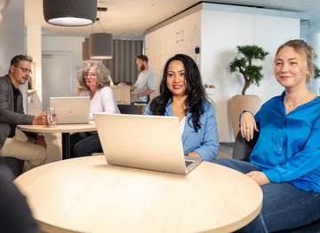 Mehrere Personen in einem modernen Büro treffen sich in entspannter Atmosphäre an runden Tischen. Zwei Frauen im Vordergrund arbeiten an Laptops, während andere im Hintergrund im Gespräch vertieft sind.