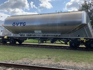 Ein Kesselwagen aus Edelstahl mit einem VTG-Logo steht auf den Gleisen, umgeben von Gras und Bäumen unter einem teilweise bewölkten Himmel.